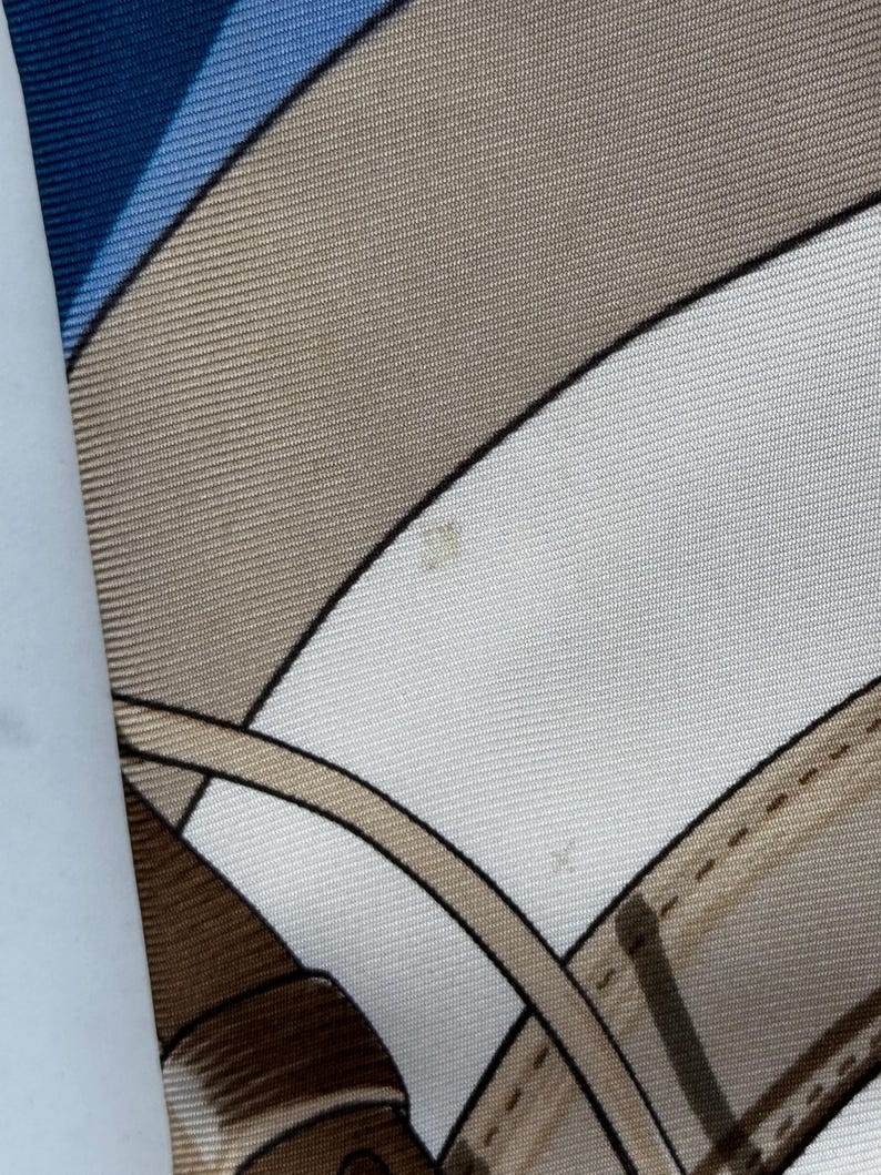 Puede incluir: Pa&ntilde;uelo de seda con un dise&ntilde;o geom&eacute;trico en tonos azul, beige y blanco. El pa&ntilde;uelo presenta l&iacute;neas curvas y un patr&oacute;n que recuerda elementos ecuestres.