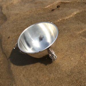 May include: A silver bowl with a shiny finish sits on a sandy surface. The bowl has three legs shaped like seahorses.