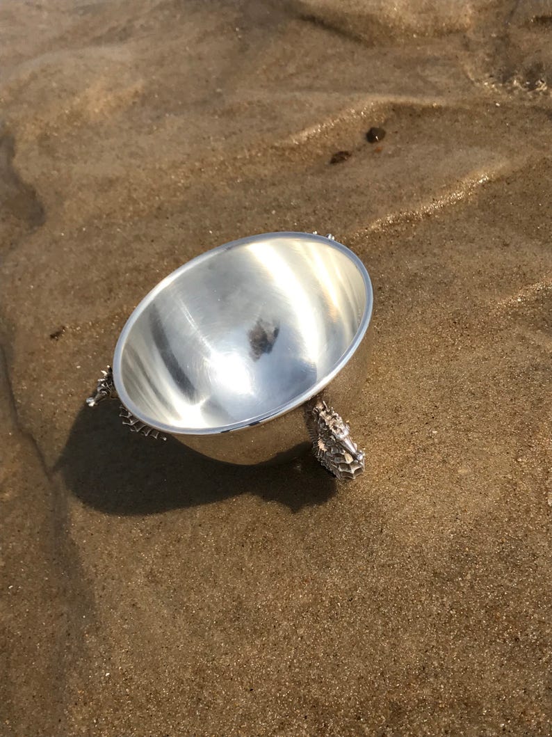 May include: A silver bowl with a shiny finish sits on a sandy surface. The bowl has three legs shaped like seahorses.