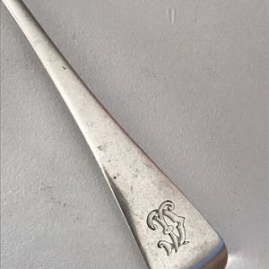 Może przedstawiać: Zbliżenie srebrnej łyżki z monogramem na trzonku. Łyżka ma prostokątny trzonek z zaokrąglonymi krawędziami. Monogram to ozdobny wz&oacute;r splecionych liter.