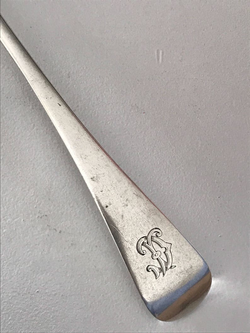 Może przedstawiać: Zbliżenie srebrnej łyżki z monogramem na trzonku. Łyżka ma prostokątny trzonek z zaokrąglonymi krawędziami. Monogram to ozdobny wz&oacute;r splecionych liter.