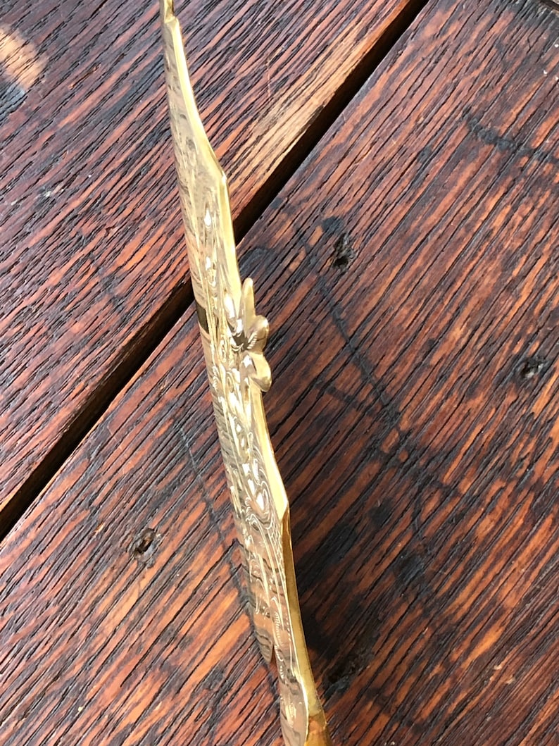 May include: A gold-colored metal decorative wall plaque with an ornate design. The plaque has a floral pattern and a raised edge.