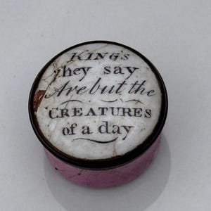 May include: A small, round, antique trinket box with a pink base and a white lid. The lid features the text "Kings they say Are but the CREATURES of a day" in black script. The box has a dark brown rim.