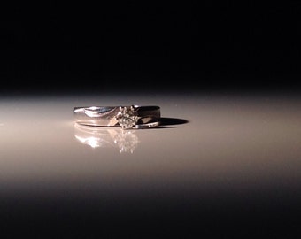 May include: A silver ring with a single diamond set in the center. The ring is on a white surface.