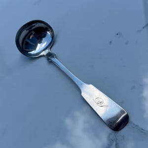 May include: A silver ladle with a round bowl and a long handle. The handle has a rectangular end with an engraved design. The ladle is reflective, showing the surrounding environment. A classic kitchen utensil.