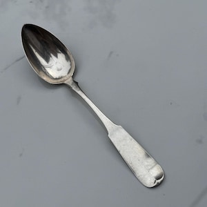 May include: A silver spoon with an oval bowl and a long, slender handle. The spoon has a simple, elegant design, and the metal has a polished, reflective surface. The spoon is likely vintage or antique.