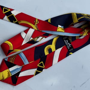 Puede incluir: Una corbata de seda roja, azul marino y dorada con un patr&oacute;n repetido de elementos ecuestres. La corbata presenta cadenas doradas, hebillas y rayas blancas. La marca "HERMES - PARIS" es visible en una etiqueta.