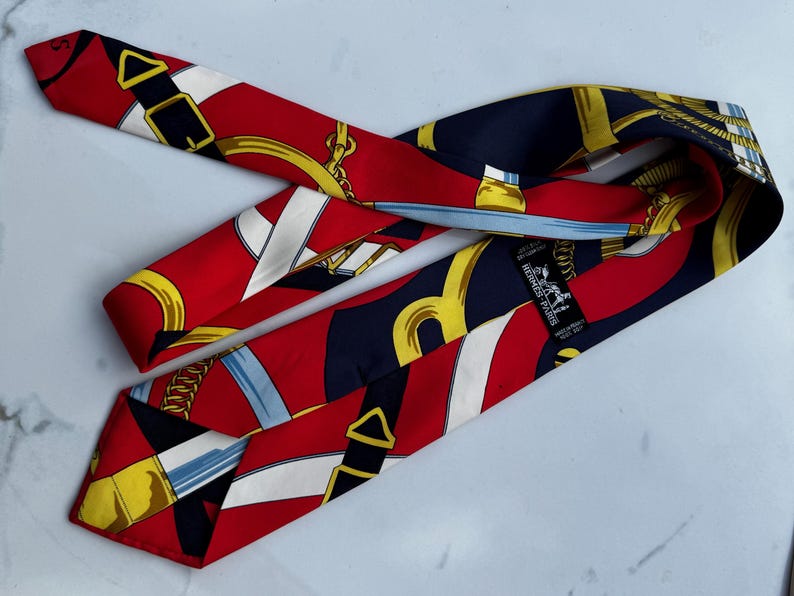 Puede incluir: Una corbata de seda roja, azul marino y dorada con un patr&oacute;n repetido de elementos ecuestres. La corbata presenta cadenas doradas, hebillas y rayas blancas. La marca "HERMES - PARIS" es visible en una etiqueta.