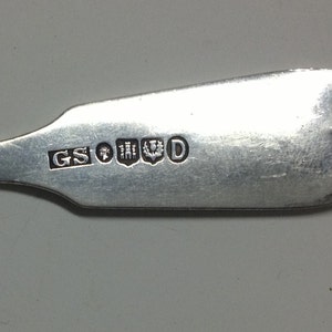 Puede incluir: Primer plano de la parte posterior de una cuchara de plata con los punzones "GS" y "D" y un escudo de armas.