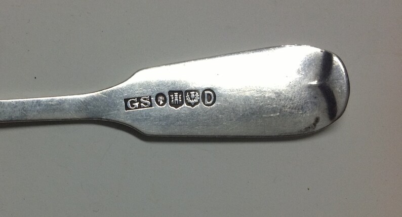 Puede incluir: Primer plano de la parte posterior de una cuchara de plata con los punzones "GS" y "D" y un escudo de armas.