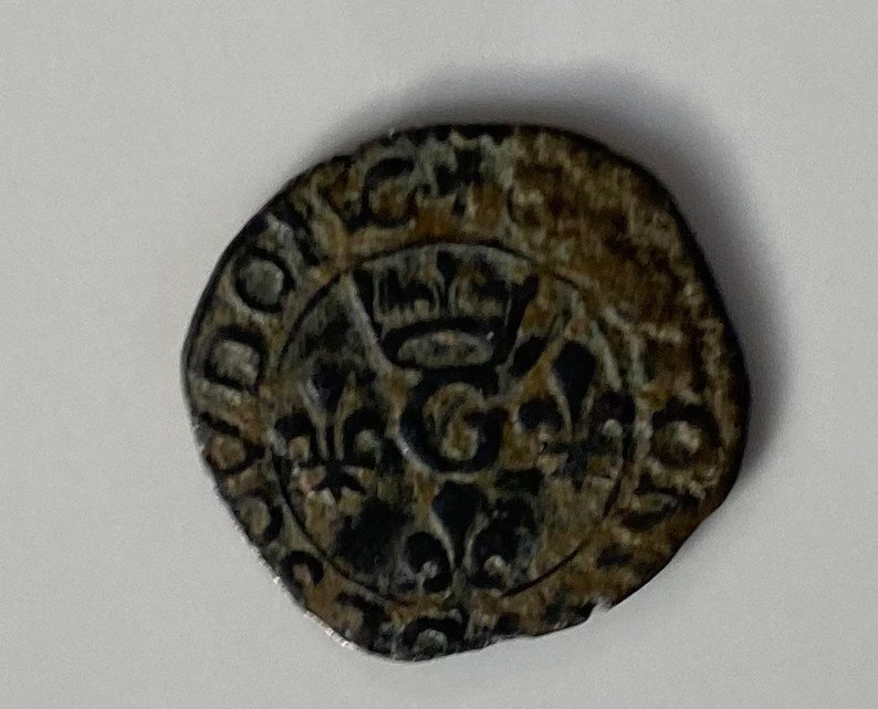 May include: A silver coin with a crown and a letter G in the centre. The coin is surrounded by a circular inscription.