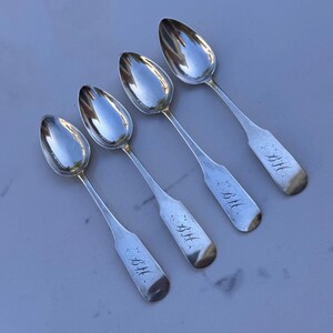 May include: Four antique silver spoons with oval bowls and long, slender handles. Each spoon has a rounded end and is engraved with the initials "B.M.". The spoons are arranged diagonally on a light-colored surface, reflecting the light.