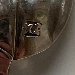 May include: Close-up of a silver spoon with the mark "XE" stamped on the back.