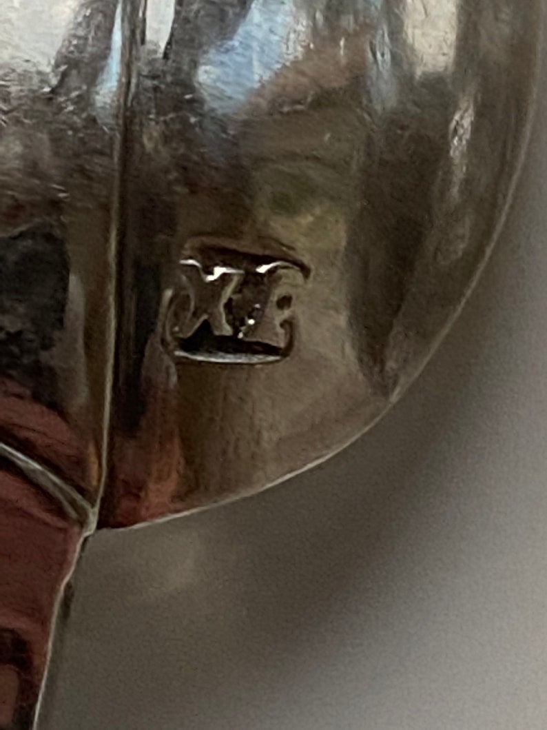 May include: Close-up of a silver spoon with the mark "XE" stamped on the back.