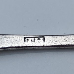 K&ouml;nnte beinhalten: Nahaufnahme eines silbernen L&ouml;ffels mit einer Punze, die "TUT" in einer rechteckigen Form zeigt.