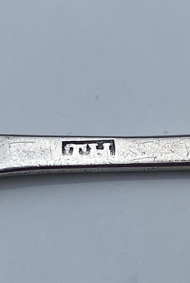 K&ouml;nnte beinhalten: Nahaufnahme eines silbernen L&ouml;ffels mit einer Punze, die "TUT" in einer rechteckigen Form zeigt.