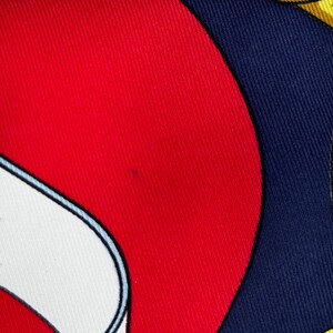 Puede incluir: Primer plano de un pa&ntilde;uelo de seda con un dise&ntilde;o audaz. El pa&ntilde;uelo presenta una combinaci&oacute;n de colores rojo vibrante, blanco y azul marino, con un patr&oacute;n amarillo en forma de cadena. La tela tiene un brillo sutil, lo que sugiere un material lujoso.