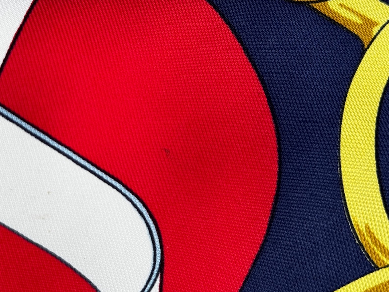 Puede incluir: Primer plano de un pa&ntilde;uelo de seda con un dise&ntilde;o audaz. El pa&ntilde;uelo presenta una combinaci&oacute;n de colores rojo vibrante, blanco y azul marino, con un patr&oacute;n amarillo en forma de cadena. La tela tiene un brillo sutil, lo que sugiere un material lujoso.