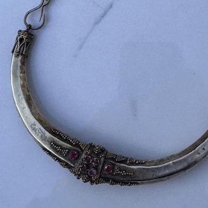 May include: A crescent-shaped silver-toned necklace featuring a hammered metal finish, ornate decorative end caps, and a central cluster of small, round, deep red stones set within intricate, textured metalwork attached to a thin metal chain.