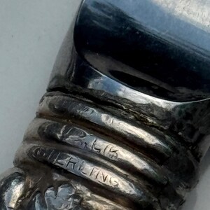 May include: Close-up of a silver object with the word "STERLING" engraved. The object has a reflective, curved surface and a textured, ribbed design. The base of the object has a floral pattern.