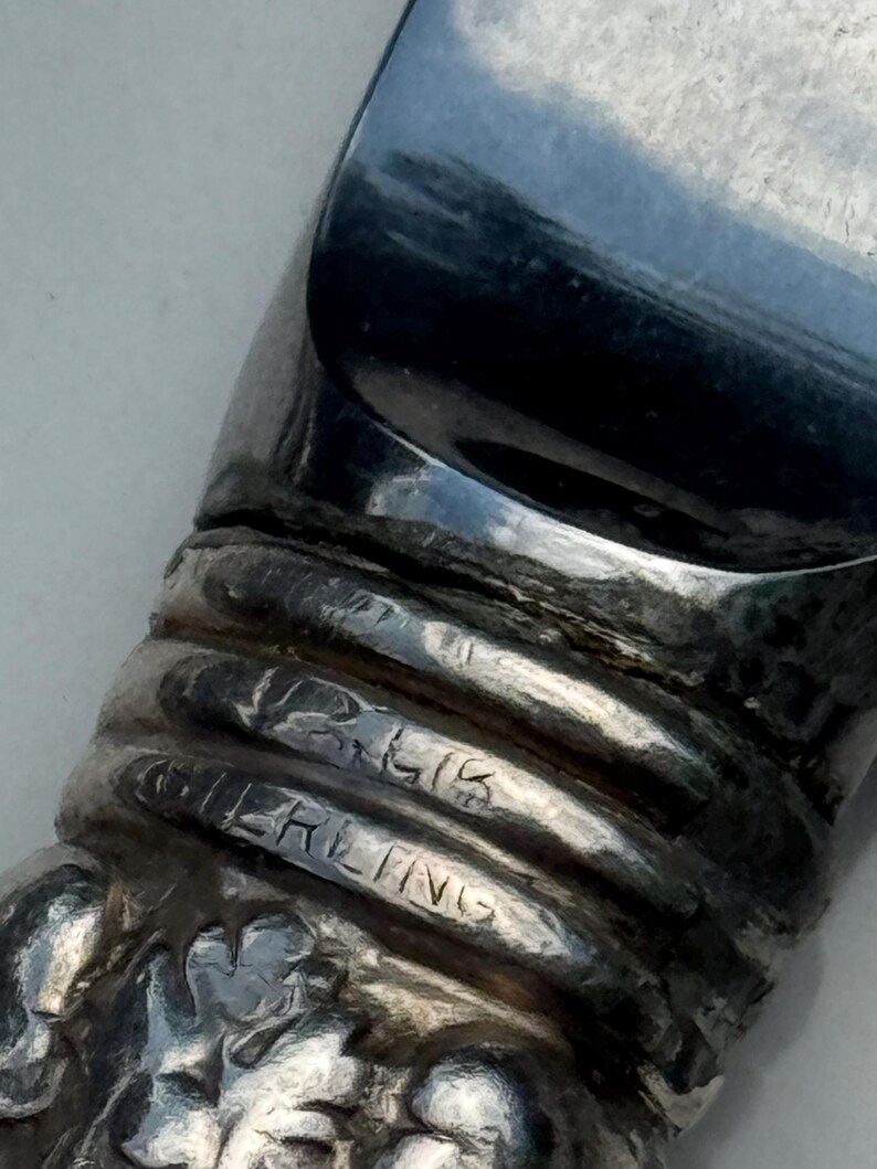 May include: Close-up of a silver object with the word "STERLING" engraved. The object has a reflective, curved surface and a textured, ribbed design. The base of the object has a floral pattern.