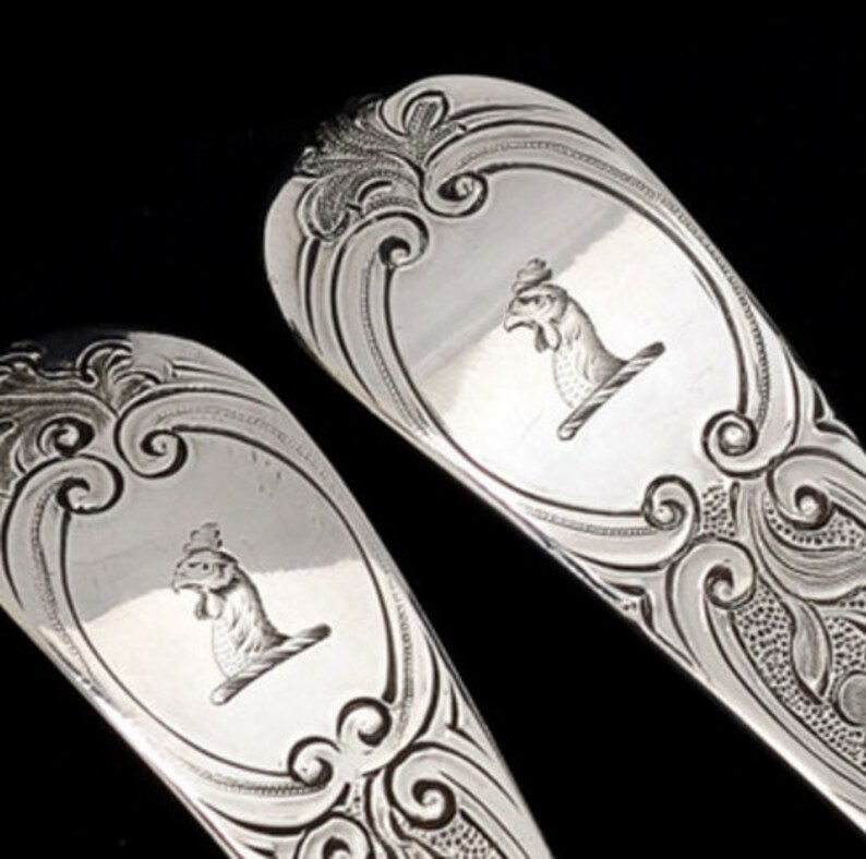 May include: Close-up of two ornate silver spoons. Each spoon features a detailed engraved design with a central oval containing a bird motif. The spoons have decorative scrollwork and a polished finish, set against a black background.
