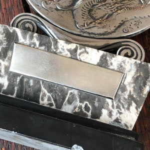 May include: A silver-toned decorative dish with a raised design of a fish. The dish is mounted on a black and white marble base with a silver-toned rectangular plaque.