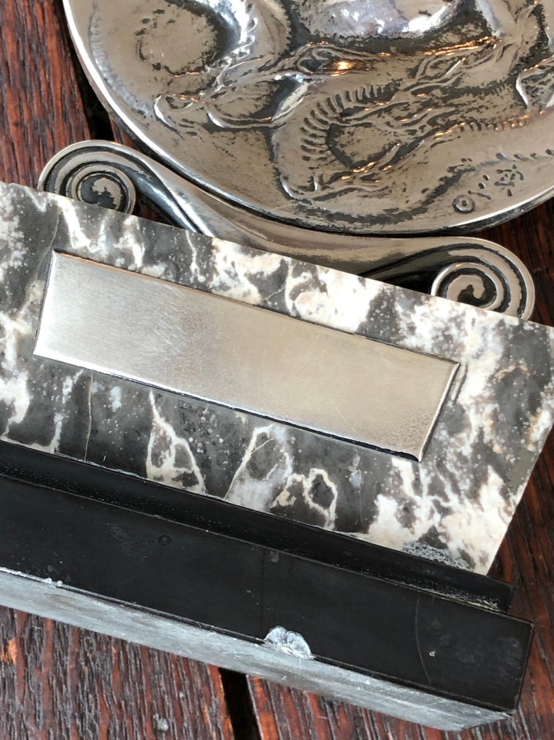 May include: A silver-toned decorative dish with a raised design of a fish. The dish is mounted on a black and white marble base with a silver-toned rectangular plaque.