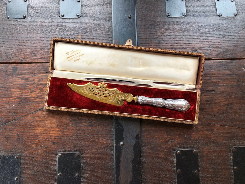 May include: A silver and gold cake knife with an ornate design, in a red velvet lined box. The box is labeled "H. Williams, Made in France, London".