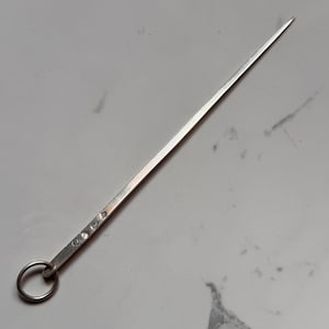 May include: A long, slender, silver-toned metal skewer or pick featuring a pointed tip and a circular ring attached to the flat, square-edged handle. The handle displays a series of small, stamped hallmarks along its side, indicating metal quality.