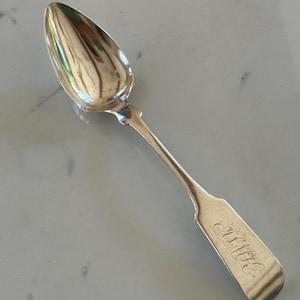 May include: A silver spoon with an oval bowl and a long, slender handle. The handle has a decorative element near the bowl and a rectangular end with engraved initials. The spoon is reflective, showing the surrounding environment.