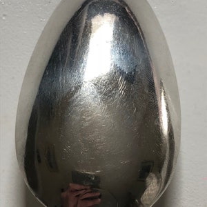 Puede incluir: Una cuchara de plata con una superficie brillante y reflectante. La cuchara tiene forma ovalada y un cuenco redondeado.