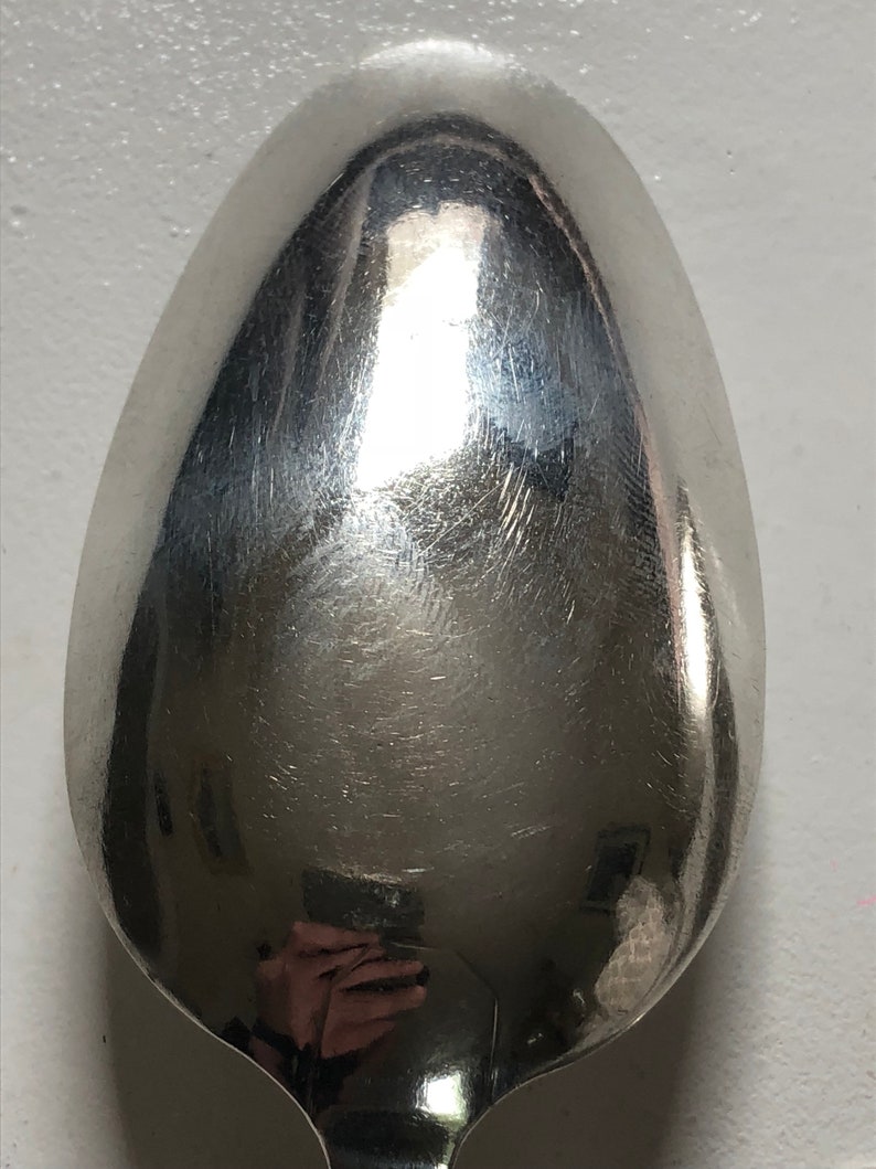 Puede incluir: Una cuchara de plata con una superficie brillante y reflectante. La cuchara tiene forma ovalada y un cuenco redondeado.