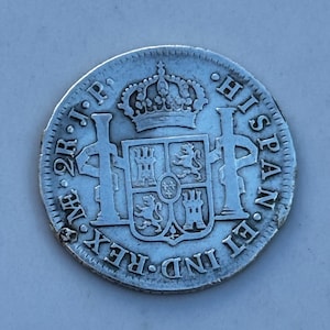 K&ouml;nnte beinhalten: Eine silberne M&uuml;nze mit aufwendigen Details, die eine k&ouml;nigliche Krone &uuml;ber einem Wappen zeigt. Der Rand der M&uuml;nze ist strukturiert, und die Inschrift "HISPAN ET IND REX" ist sichtbar. Die M&uuml;nze scheint antik zu sein.