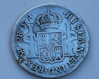 May include: A silver coin with intricate details, featuring a royal crown above a coat of arms. The coin's edge is textured, and the inscription "HISPAN ET IND REX" is visible. The coin appears to be an antique, with signs of wear.