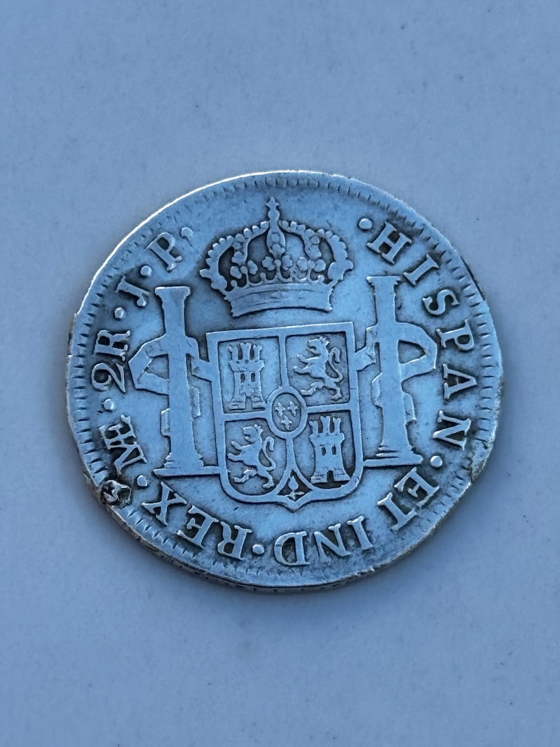 K&ouml;nnte beinhalten: Eine silberne M&uuml;nze mit aufwendigen Details, die eine k&ouml;nigliche Krone &uuml;ber einem Wappen zeigt. Der Rand der M&uuml;nze ist strukturiert, und die Inschrift "HISPAN ET IND REX" ist sichtbar. Die M&uuml;nze scheint antik zu sein.