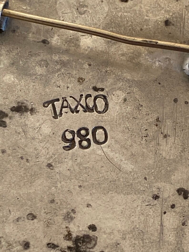 May include: Close-up of a silver pin with the words "Taxco 980" stamped on the back.