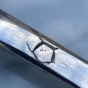 May include: Close-up of a silver metal object with a geometric mark. The mark is a stylized shape with a diamond in the center.