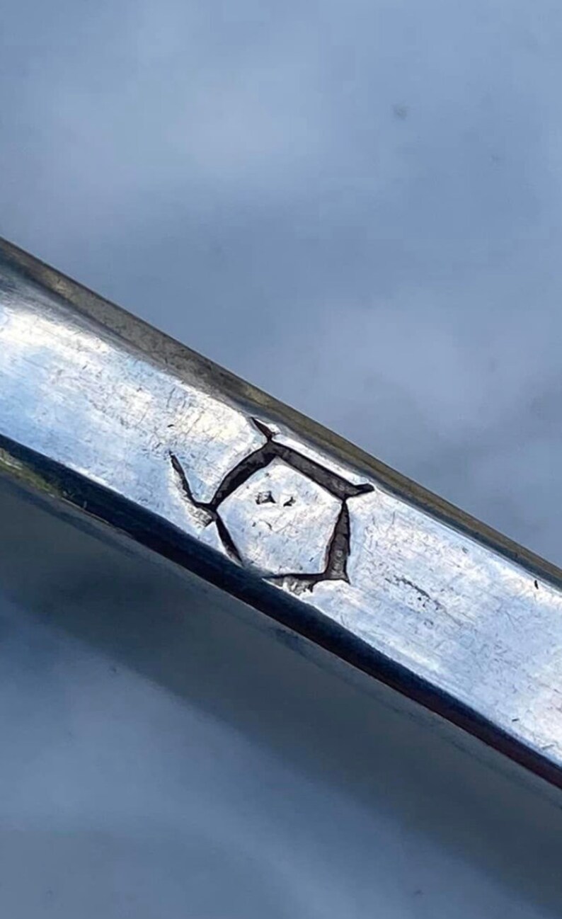 May include: Close-up of a silver metal object with a geometric mark. The mark is a stylized shape with a diamond in the center.