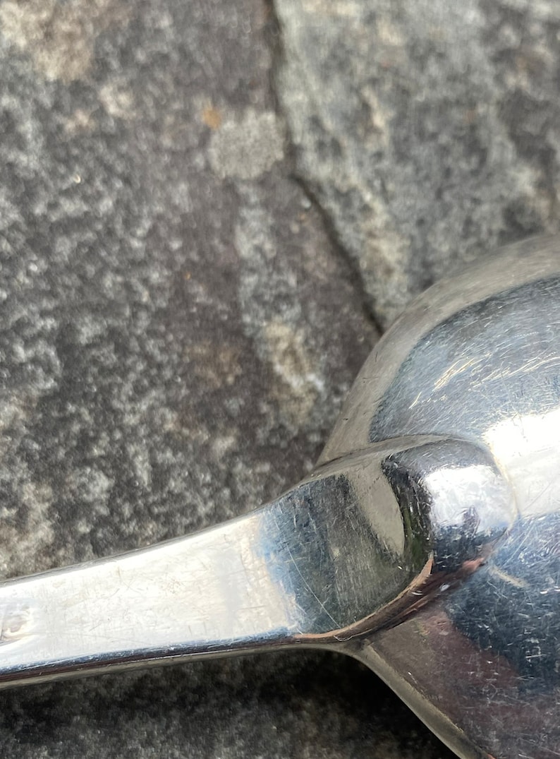 May include: Close-up of a silver spoon with a rounded bowl and a long, slender handle. The spoon is lying on a rough, gray surface.