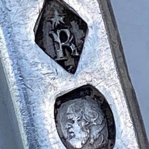 May include: Close-up of a silver hallmark with a diamond-shaped mark containing the letter "R" and a smaller mark with a crowned head.