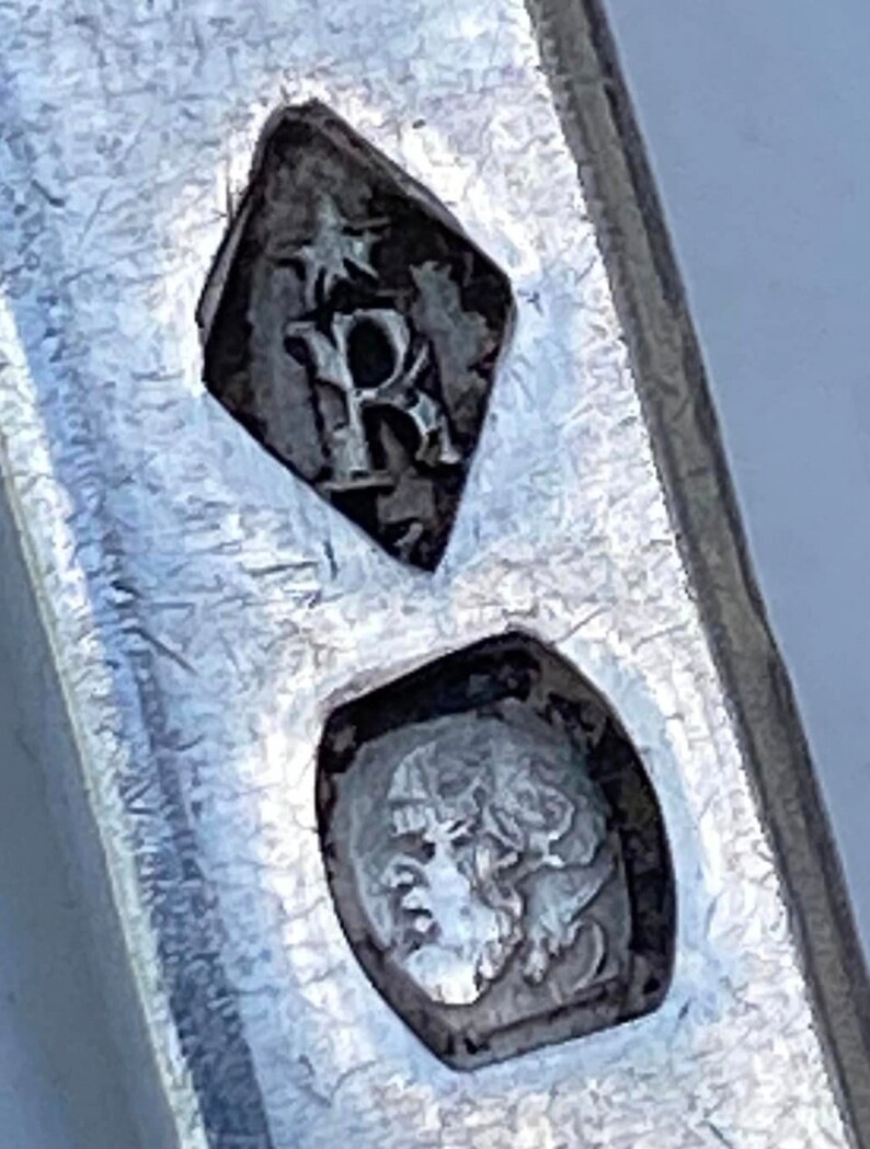 May include: Close-up of a silver hallmark with a diamond-shaped mark containing the letter "R" and a smaller mark with a crowned head.