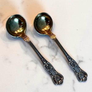 May include: Two antique silver spoons with gold-coloured bowls and ornate handles. The spoons are displayed on a white surface, showcasing their intricate design and reflective surfaces. The spoons are likely for serving.