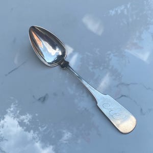 May include: A silver spoon with a rounded, pointed bowl and a long, slender handle. The handle is engraved with the initials "C.C."