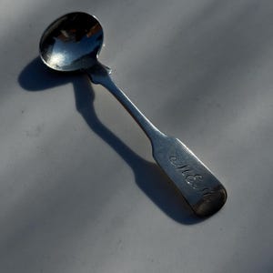 May include: A small, silver spoon with a rounded bowl and a long, slender handle. The handle has an engraved monogram. The spoon is likely vintage or antique, and is resting on a light gray surface.