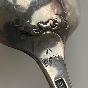 K&ouml;nnte beinhalten: Nahaufnahme eines silbernen L&ouml;ffels mit einem verzierten Design auf der R&uuml;ckseite. Der L&ouml;ffel tr&auml;gt die Initialen "R.H." und "M" auf dem Griff.
