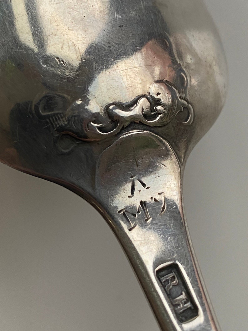K&ouml;nnte beinhalten: Nahaufnahme eines silbernen L&ouml;ffels mit einem verzierten Design auf der R&uuml;ckseite. Der L&ouml;ffel tr&auml;gt die Initialen "R.H." und "M" auf dem Griff.