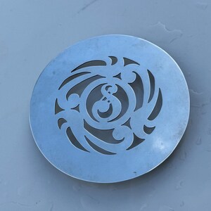 May include: A silver-coloured metal coaster with a decorative cut-out design featuring a stylised swirl pattern and a central letter "S".