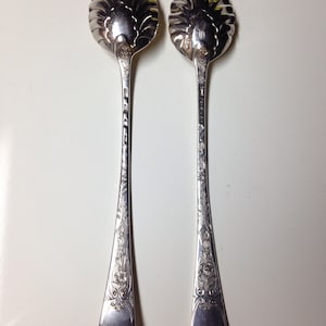 May include: Two silver serving spoons with ornate floral and scrollwork designs. The spoon bowls have a scalloped edge. The handles are long and slender, with detailed engravings. The spoons are displayed on a white surface.