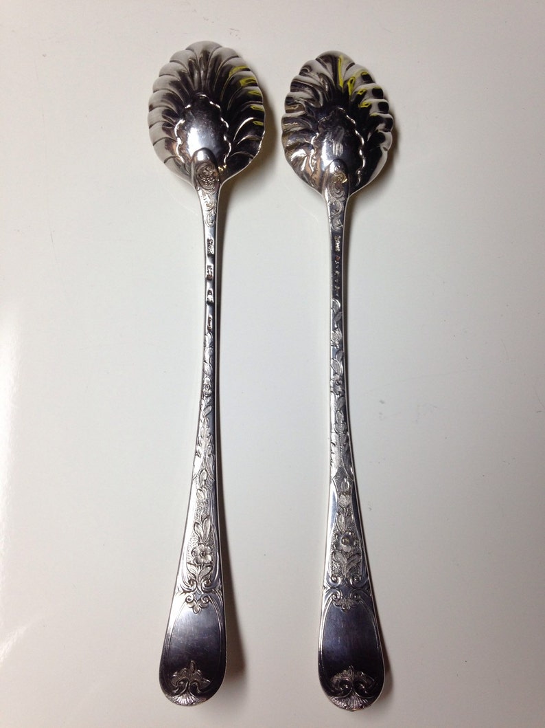 May include: Two silver serving spoons with ornate floral and scrollwork designs. The spoon bowls have a scalloped edge. The handles are long and slender, with detailed engravings. The spoons are displayed on a white surface.